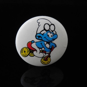 Könnte beinhalten: Ein weißer Knopf mit einem blauen Schlumpf, der ein rotes Dreirad mit gelben Rädern fährt. Der Schlumpf trägt einen weißen Hut und hat ein blaues Gesicht mit einem roten Mund.