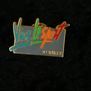 Op de afbeelding: Een gouden pin met een grijze achtergrond en de tekst "Vive le Sport" in kleurrijke letters. De tekst "Renault" is in kleinere letters onder de hoofdtekst gedrukt.
