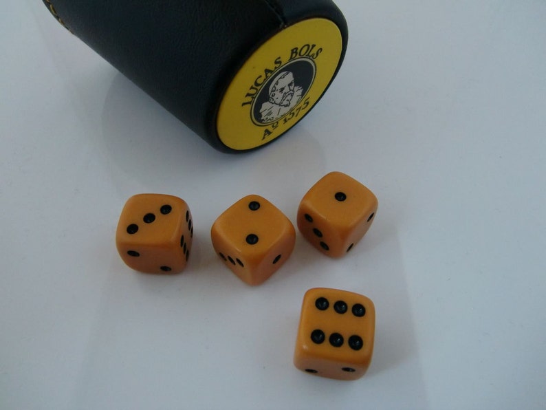 May include: Four orange dice with black dots on a white surface. The dice are next to a black and yellow dice cup with the text "Lucas Bols 1575" on it.