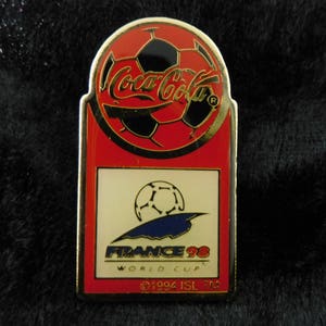 Può includere: Uno spillo Coca-Cola rosso, bianco e oro con un pallone da calcio con il testo "France 98 World Cup" e "©1994 ISL TM".