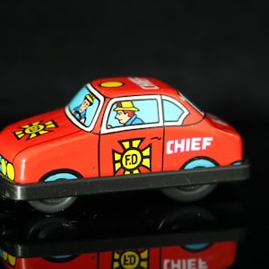 Könnte beinhalten: Rotes Spielzeugfeuerwehrauto mit schwarzer Basis und blauen Rädern. Das Auto hat einen gelben Stern mit den Buchstaben "F.D." darin und das Wort "CHIEF" an der Seite. Zwei Comic-Feuerwehrleute sind im Auto zu sehen.