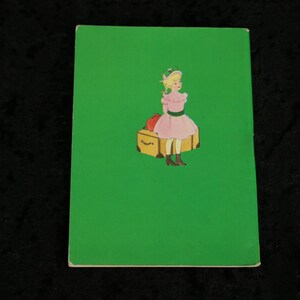 Puede incluir: Un libro verde con una imagen de una ni&ntilde;a con un vestido rosa sentada en una maleta con una manzana roja encima. La ni&ntilde;a lleva un sombrero verde y tiene el pelo rubio.