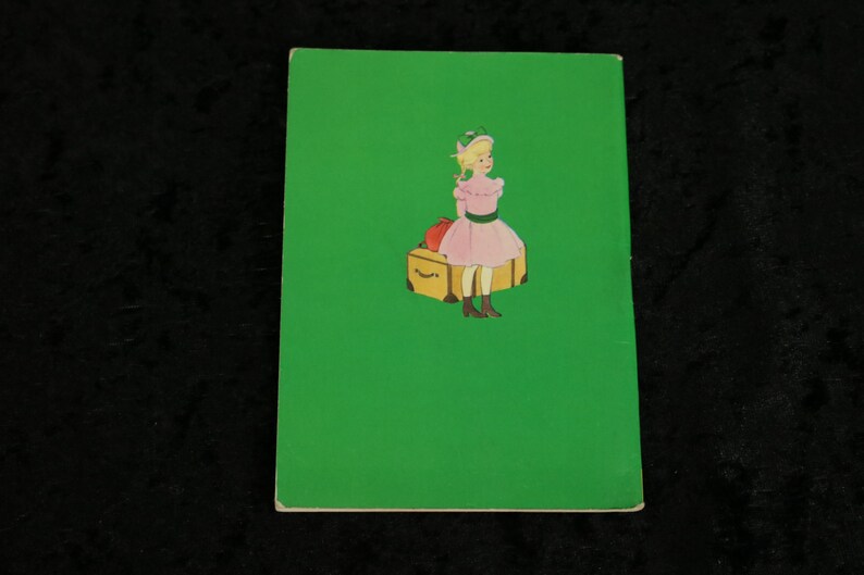 Puede incluir: Un libro verde con una imagen de una ni&ntilde;a con un vestido rosa sentada en una maleta con una manzana roja encima. La ni&ntilde;a lleva un sombrero verde y tiene el pelo rubio.