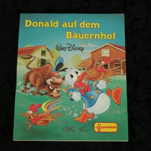 Peut inclure: Une couverture de livre pour enfants vintage mettant en scène Donald Duck dans une ferme. La couverture est bleue et verte avec des illustrations de Donald Duck, un taureau et un coq. Le titre du livre est "Donald auf dem Bauernhof" et le livre est publié par Walt Disney.