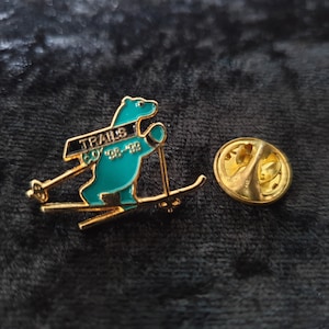 Könnte beinhalten: Goldfarbene Anstecknadel mit einem grünen Bären, der Ski fährt, und dem Text "Trails CO '98-'99".