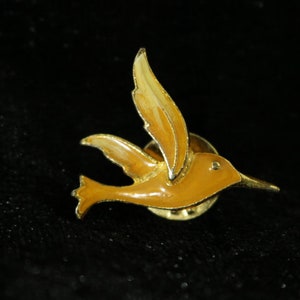Könnte beinhalten: Eine goldfarbene Kolibri-Brosche mit einer tanfarbenen Emaille-Oberfläche. Der Vogel wird im Flug mit ausgebreiteten Flügeln dargestellt. Die Brosche hat ein klassisches Design und eignet sich für Jacken, Hüte oder Taschen.