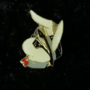 Könnte beinhalten: Eine goldfarbene Anstecknadel mit einer weißen Comicfigur, die einen schwarzen Hut und eine Sonnenbrille trägt. Die Figur hat einen rot-weißen Schal um den Hals.