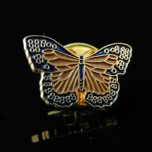 Könnte beinhalten: Eine goldfarbene Schmetterlingsbrosche mit aufwendigen Details. Die Flügel zeigen eine Kombination aus braunem und blauem Emaille, umrandet mit Gold. Die Brosche ist in einer klassischen Schmetterlingsform gestaltet, perfekt für einen Hauch von Natur.