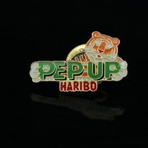Könnte beinhalten: Eine grüne und weiße Emaille-Anstecknadel mit den Worten "PEP-UP HARIBO" und einem Comic-Tiger.