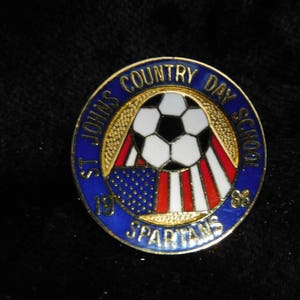 Peut inclure: Une épingle bleue et dorée avec les mots "St. Johns Country Day School" et "Spartans" autour d'un ballon de football avec un motif de drapeau américain. L'épingle porte également les années "79" et "88".