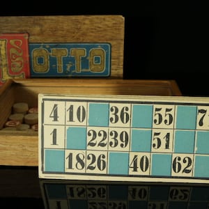 Könnte beinhalten: Eine antike Holzkiste mit dem Wort "LOTTO" auf dem Deckel. Die Kiste enthält einen Satz hölzerner Bingokugeln und eine blaue und weiße Bingokarte mit den Zahlen 4, 10, 36, 55, 71, 1, 22, 39, 51, 18, 26, 40, 62, 86 und 88.