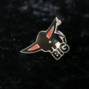 Könnte beinhalten: Eine schwarze und rote Emaille-Anstecknadel mit einem Cartoon-Stierkopf und dem Wort "BIG" in weißen Buchstaben.