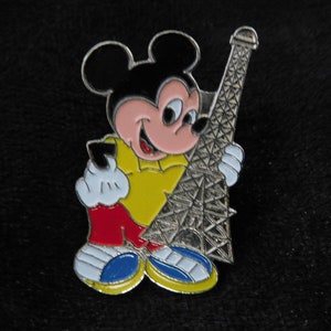 Könnte beinhalten: Eine emaillierte Anstecknadel mit Mickey Mouse, der den Eiffelturm hält. Mickey trägt ein gelbes Hemd, rote Hosen und blaue Schuhe. Der Eiffelturm ist silberfarben.