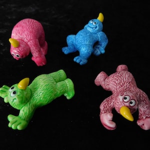 Könnte beinhalten: Vier kleine Plüschtiere in Form von Monstern. Die Monster sind rosa, blau, grün und rot. Jedes Monster hat einen anderen Gesichtsausdruck und ein gelbes Horn auf dem Kopf.