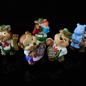May include: A set of seven small figurines, including bears, a hippopotamus, and a girl. The bears are wearing traditional German clothing and holding various items, such as a microphone, a heart-shaped cookie, and a sign that says "Ich liebe Dich". The hippopotamus is blue and wearing a red and white striped shirt. The girl is wearing a blue dress and holding a red and white striped bag.