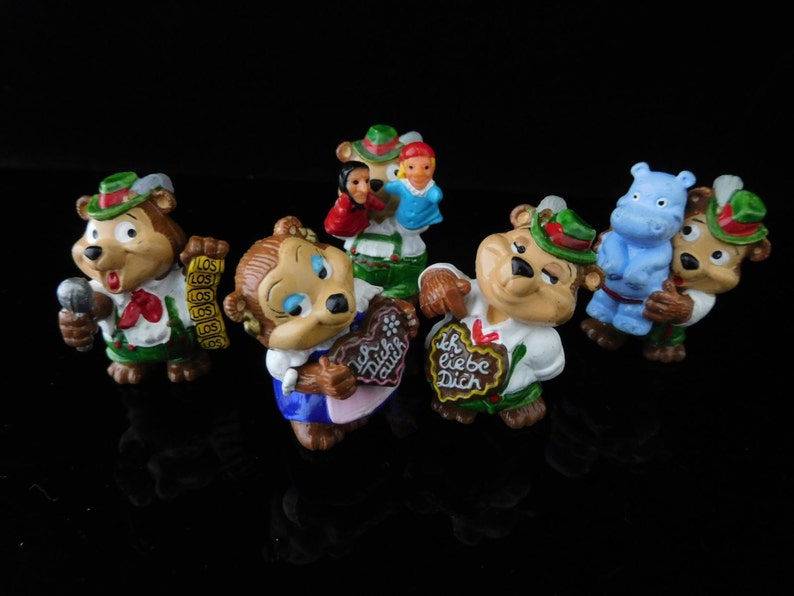 May include: A set of seven small figurines, including bears, a hippopotamus, and a girl. The bears are wearing traditional German clothing and holding various items, such as a microphone, a heart-shaped cookie, and a sign that says "Ich liebe Dich". The hippopotamus is blue and wearing a red and white striped shirt. The girl is wearing a blue dress and holding a red and white striped bag.