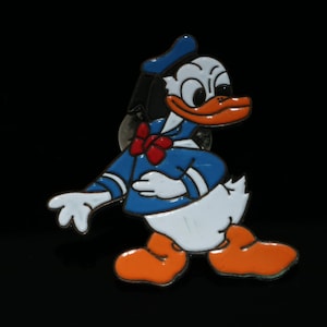 Könnte beinhalten: Eine Cartoon-Anstecknadel von Donald Duck, der ein blaues Matrosenhemd mit einer roten Schleife und einer blauen Matrosenmütze trägt. Er hat einen weißen Körper mit orangenen Füßen und einen gelben Schnabel.
