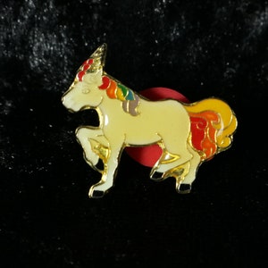 Könnte beinhalten: Eine goldfarbene Einhorn-Anstecknadel mit weißem Körper, regenbogenfarbener Mähne und Schwanz und roter Basis.