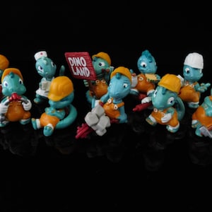 Könnte beinhalten: Eine Sammlung von zehn kleinen blauen und gelben Dinosaurier-Figuren, die Bauarbeiterhüte tragen und Werkzeuge halten. Die Figuren sind auf einer schwarzen Oberfläche angeordnet. Ein Schild lautet "Dino Land".