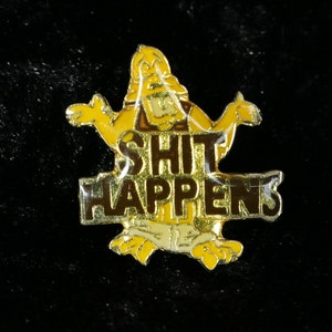 Könnte beinhalten: Eine goldfarbene Anstecknadel mit einer Comicfigur, die ein Schild mit der Aufschrift "SHIT HAPPENS" hält.