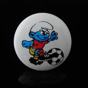 May include: A white button with a cartoon image of a blue Smurf playing soccer. The Smurf is wearing a red and white jersey and blue shorts. The Smurf is kicking a black and white soccer ball.