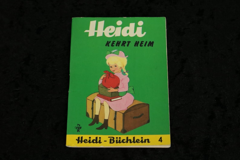 Puede incluir: Una portada de libro infantil vintage con una ni&ntilde;a de pelo rubio y un vestido rosa sentada en un cofre de madera. El t&iacute;tulo del libro es "Heidi Kehrt Heim" y el subt&iacute;tulo es "Heidi - B&uuml;chlein 4".