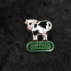 Könnte beinhalten: Eine schwarze und weiße Kuh-Anstecknadel mit grünem Hintergrund. Die Nadel trägt die Aufschrift "Westfalia Separator".