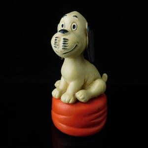 May include: A white and brown dog figurine with black ears sitting on a red and orange cushion.