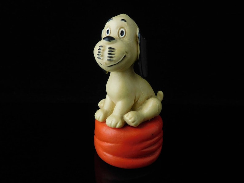 May include: A white and brown dog figurine with black ears sitting on a red and orange cushion.