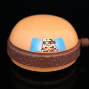 Könnte beinhalten: Ein braunes und oranges Plastikspielzeug in Form eines Hamburgers mit einer Comicfigur auf der Vorderseite. Die Figur trägt einen schwarzen Hut und ein rotes Hemd. Das Spielzeug hat einen blauen Hintergrund mit dem Text "Wendy's" darauf.