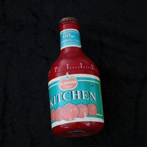 Peut inclure: Minuteur de cuisine rouge en forme de bouteille de ketchup avec une étiquette blanche indiquant "60 Minutes Timer Kitchen". L'étiquette a un fond bleu et une image de tomates rouges.
