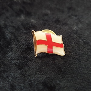 Puede incluir: Un pin de color dorado con una bandera blanca que presenta una cruz roja. La bandera es una representación de la bandera inglesa.