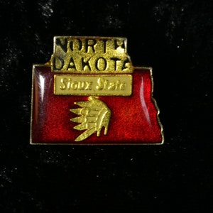 Peut inclure: Épinglette en émail rouge et or représentant l'état du Dakota du Nord avec le texte "North Dakota Sioux State" et un motif de coiffe amérindienne.