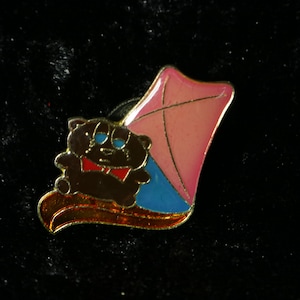 Könnte beinhalten: Eine goldfarbene Anstecknadel mit einem Comic-Bären, der eine rote Schleife trägt und auf einem blauen und rosa Drachen sitzt.