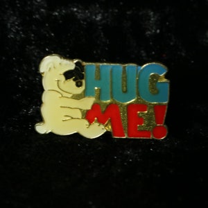 Könnte beinhalten: Ein gelber Comic-Bär mit schwarzen Augen umarmt die Buchstaben "HUG ME!" Die Buchstaben sind in einem blau-rot-goldenen Farbschema gehalten.