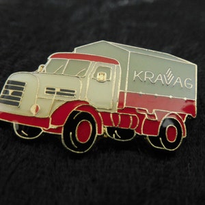 Könnte beinhalten: Eine emaillierte Anstecknadel mit einem Vintage-LKW mit einer grauen Plane. Der LKW ist weiß und rot und hat den Schriftzug "KRAVAG" auf der Seite.