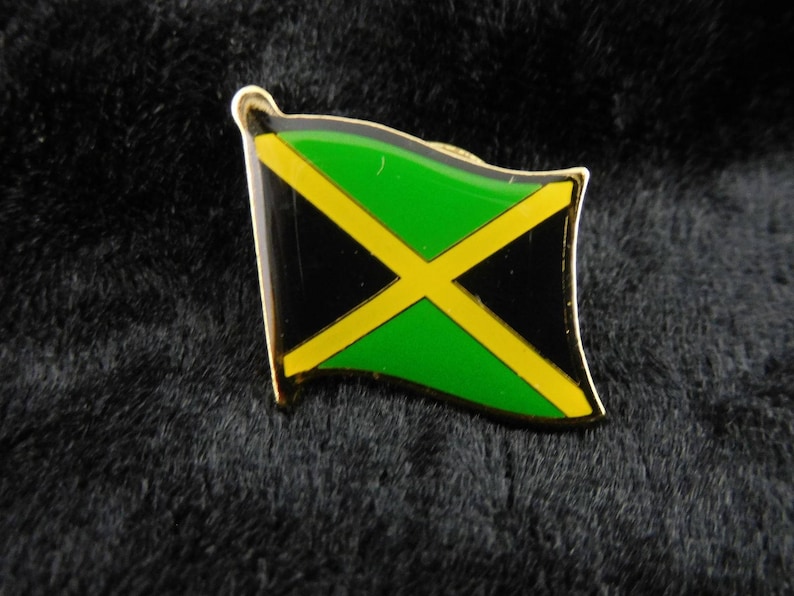 May include: A gold-coloured metal pin with a waving Jamaican flag design. The flag features a green, black, and yellow colour scheme with a yellow diagonal cross.
