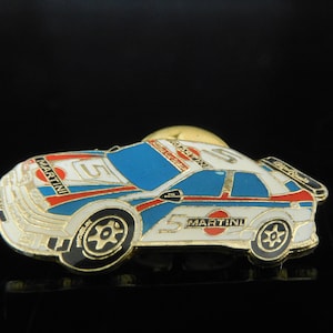 Könnte beinhalten: Eine weiße, blaue und rote Rennwagen-Anstecknadel mit der Nummer 5 und dem Martini-Logo. Die Anstecknadel ist aus Metall und hat eine goldene Oberfläche.