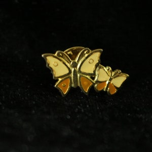 Könnte beinhalten: Eine goldfarbene Anstecknadel mit zwei Schmetterlingen. Der größere Schmetterling ist cremefarben mit orangefarbenen Akzenten. Der kleinere Schmetterling ist cremefarben mit orangefarbenen Akzenten.