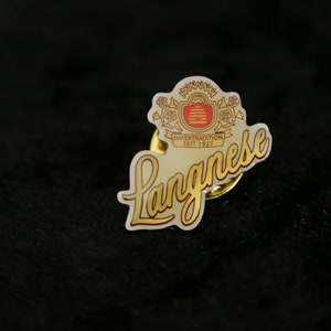 Könnte beinhalten: Eine goldene und weiße Emaille-Anstecknadel mit dem Wort "Langnese" in einer kursiv geschriebenen Schrift und einem Wappen mit dem Text "Imkertradition seit 1927".