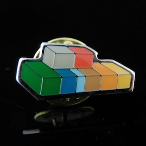 Könnte beinhalten: Eine silberne Anstecknadel mit einem farbenfrohen Blockdesign. Die Blöcke sind versetzt übereinander gestapelt und in Grün-, Blau-, Rot-, Orange- und Gelbtönen lackiert.