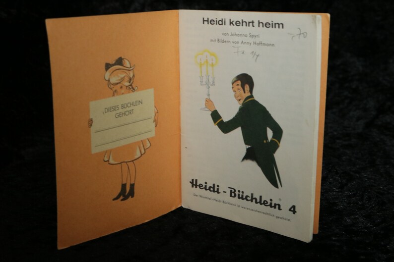 Puede incluir: Una portada de libro vintage para "Heidi kehrt heim" de Johanna Spyri, ilustrada por Anny Hoffmann. La portada presenta a una ni&ntilde;a sosteniendo un cartel que dice "DIESES BOCHLEIN GEHORT" y a un hombre con traje sosteniendo un candelabro. El libro es parte de la serie "Heidi-B&uuml;chlein", espec&iacute;ficamente el volumen 4.