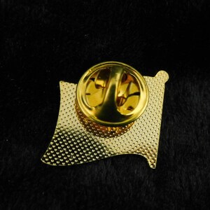May include: Gold-coloured metal pin with a textured surface and a pin back. The pin is shaped like a flag.