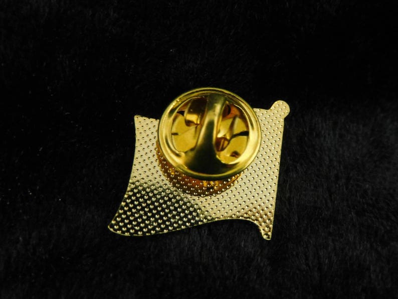 May include: Gold-coloured metal pin with a textured surface and a pin back. The pin is shaped like a flag.