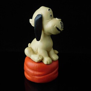 May include: A white and black dog figurine sitting on an orange cushion. The dog has a long snout and black ears.