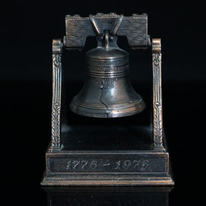 Könnte beinhalten: Eine Miniatur-Replik der Liberty Bell aus Metall mit einer dunkelbraunen Oberfläche. Die Glocke ist auf einem Sockel mit der Inschrift "1776 - 1976" montiert.