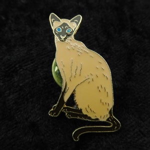 Könnte beinhalten: Eine goldfarbene Anstecknadel mit einer detaillierten Illustration einer Siamkatze mit blauen Augen. Die Katze sitzt mit ihrem Schwanz um den Körper gewickelt.