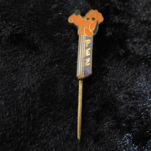 Op de afbeelding: Een gouden speld met een bruine cartoonhond die een blauwe en gouden lolly houdt met het woord "Lollipop" erop.