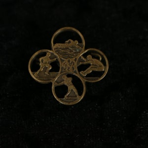 Könnte beinhalten: Eine goldfarbene Metallstifte mit vier kreisförmigen Abschnitten, die jeweils eine andere Sportart darstellen: Schwimmen, Laufen, Springen und Werfen.