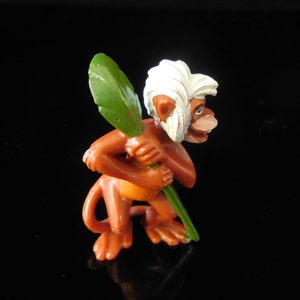 Könnte beinhalten: Eine kleine, braune Affenfigur mit weißem Fell auf dem Kopf, die ein grünes Blatt in den Händen hält. Der Affe steht auf einer schwarzen Oberfläche.